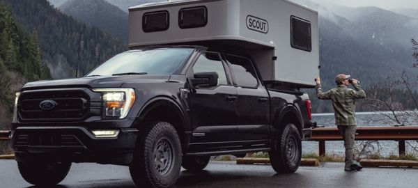 Mand drikker morgenkaffe ved bagenden af sin Truck Camper Scout fra Fraserway RV Rentals med udsigt over sø og tågede bjerge i Canada